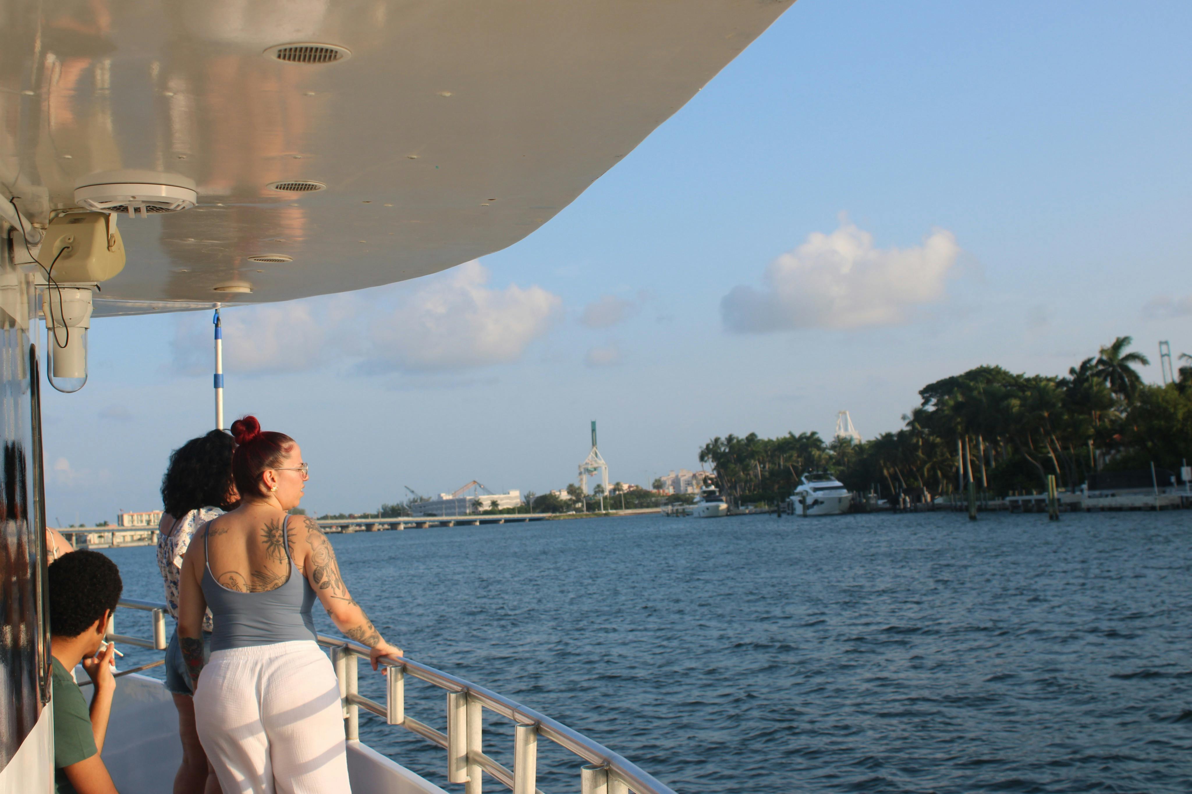 People standing and sitting on a boat's deck, enjoying a view of a calm body of water and distant buildings under a blue sky.