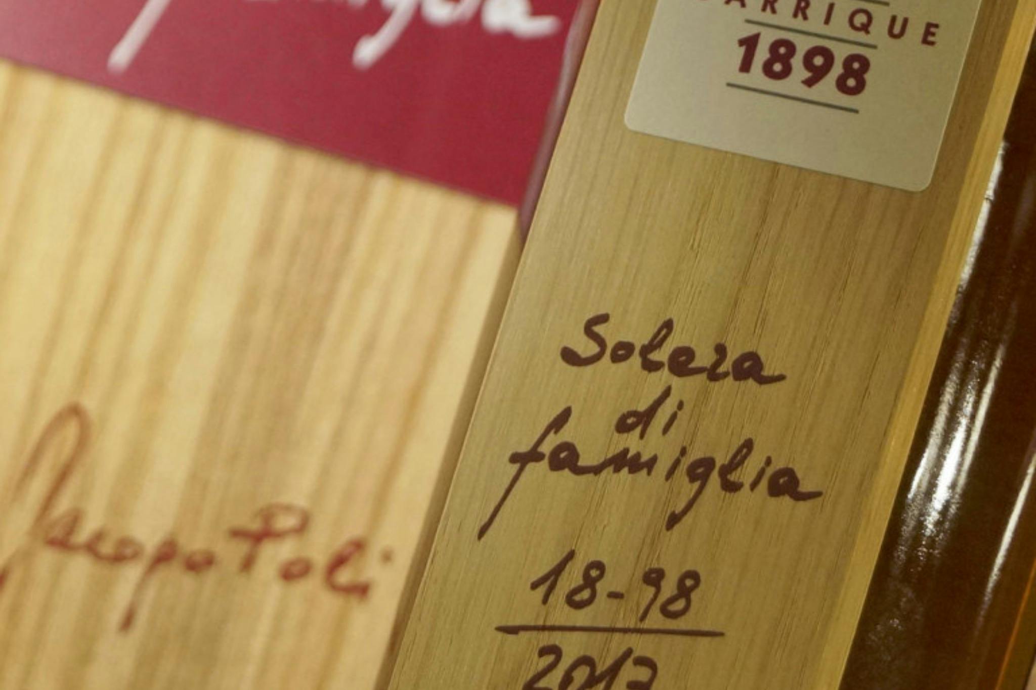 Κοντινό πλάνο ξύλινης επιφάνειας με χειρόγραφο κείμενο που γράφει «Solera di famiglia 1898-2013» και κόκκινη ετικέτα με το «1898» στην κορυφή.