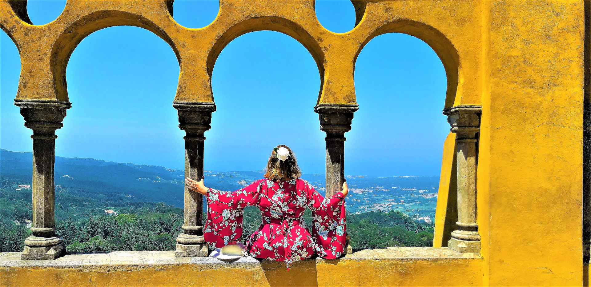 Person in red floral dress sitting between yellow arches, overlooking a scenic landscape with hills, greenery, and blue sky.