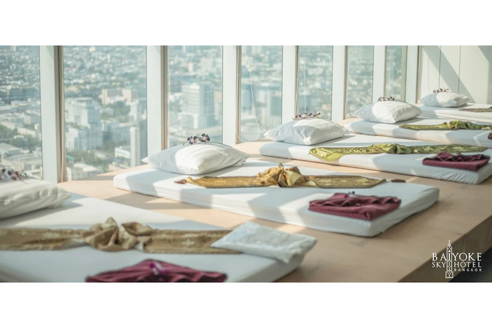 A room with neatly arranged white mattresses, each topped with a pillow and folded fabric, against large windows showing a cityscape.