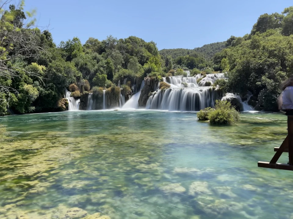 A waterfall cascading into a clear turquoise pool surrounded by lush green trees under a bright blue sky.