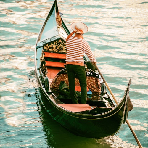 Un gondoliere con camicia e cappello a righe guida una gondola attraverso acque calme e soleggiate.