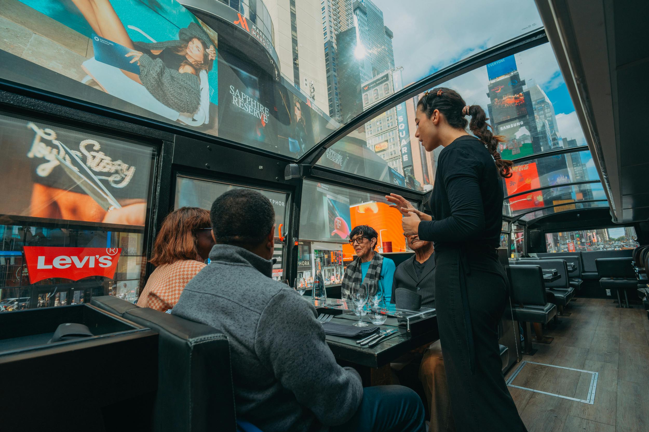 Vier mensen zitten aan een tafel in een bus met glazen dak, één vrouw staat, en buiten zijn de reclameborden van Times Square te zien.