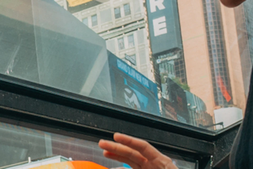 Four people sit at a table inside a glass-roofed bus, one woman standing, with Times Square billboards visible outside.