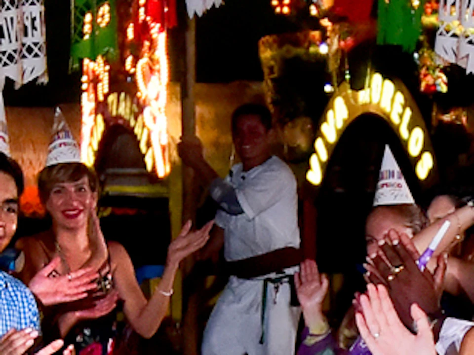 People celebrating at a long, festive table under a decorated canopy, wearing party hats, with food and drinks laid out.