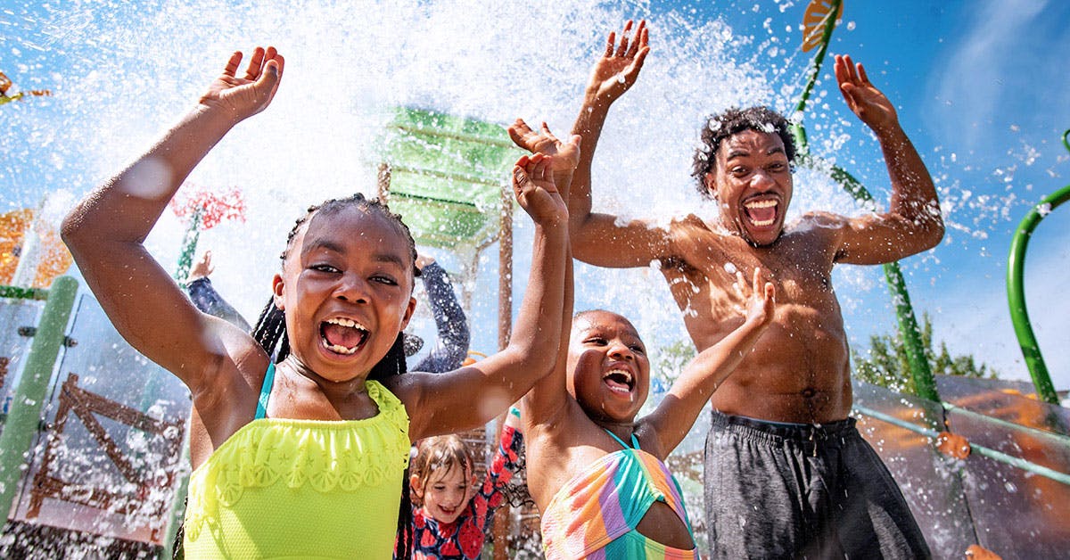Drei Kinder und ein Mann, alle in Badeanzügen, heben freudig die Arme, als das Wasser in einem Wasserpark über sie hinwegspritzt.