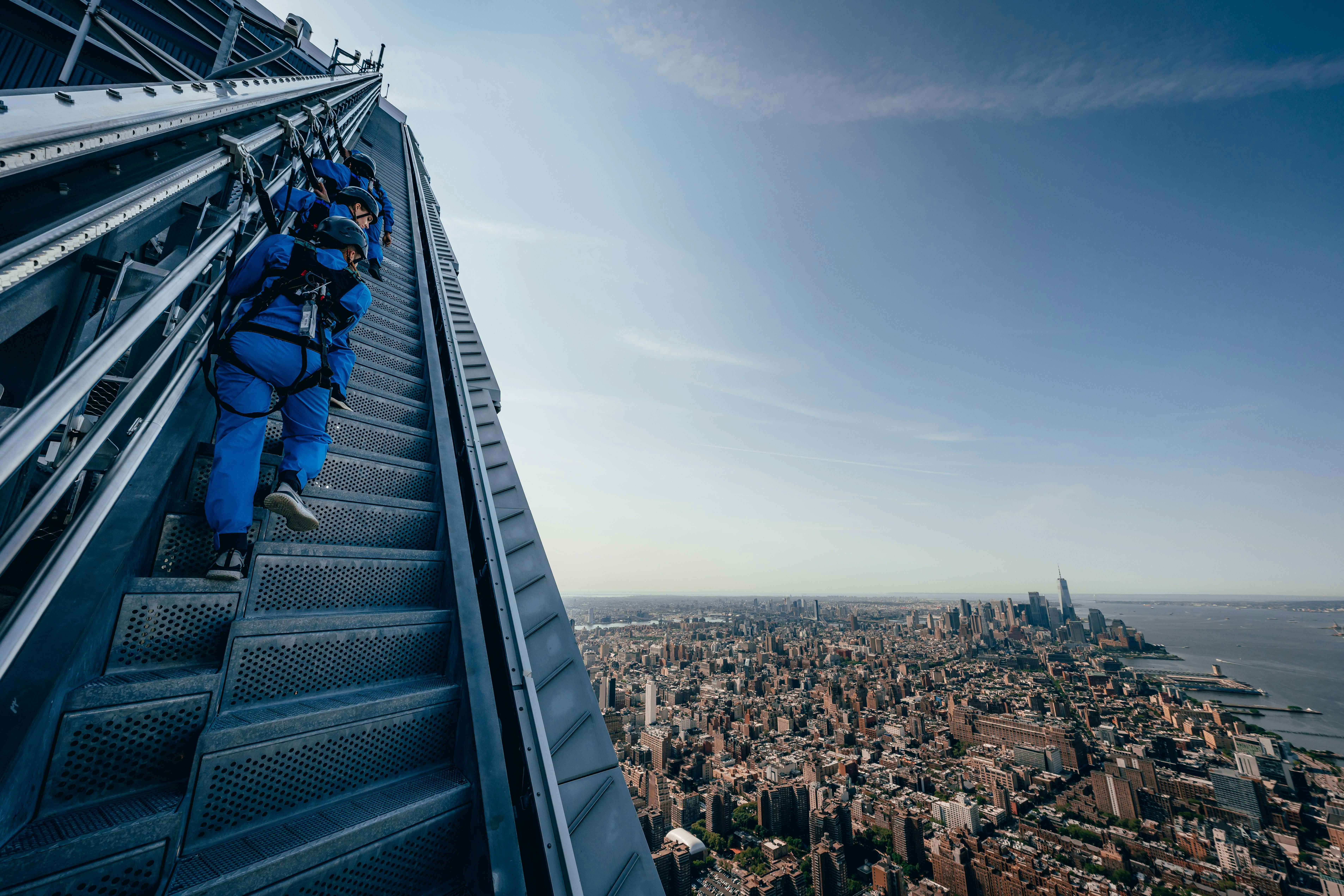 Des personnes en tenue bleue grimpent des escaliers extérieurs sur un gratte-ciel avec un paysage urbain qui s'étend en contrebas sous un ciel bleu et clair.
