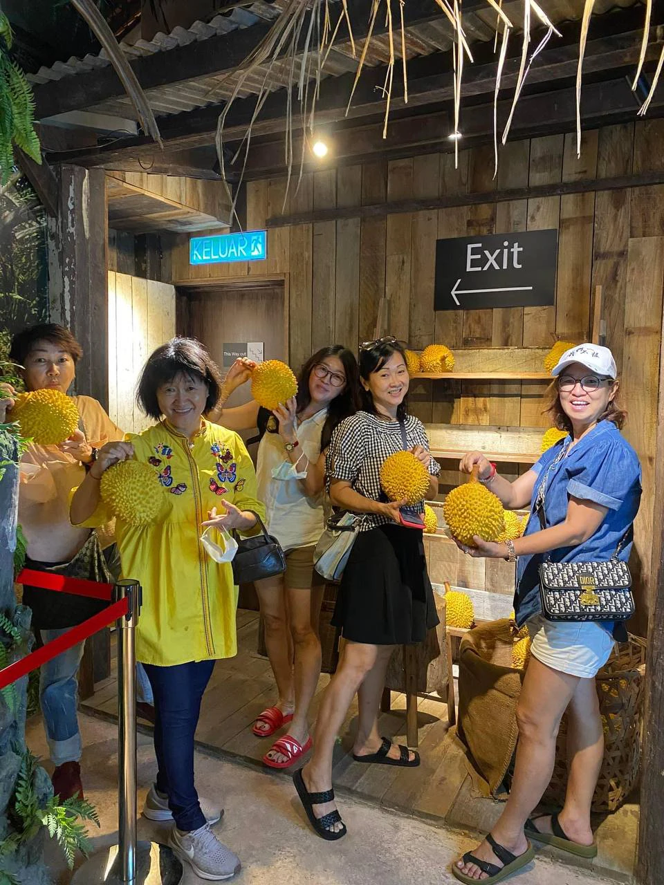 Five people holding durians inside a wooden-themed room with an 'Exit' sign and a 'Keluar' sign visible on the wall.