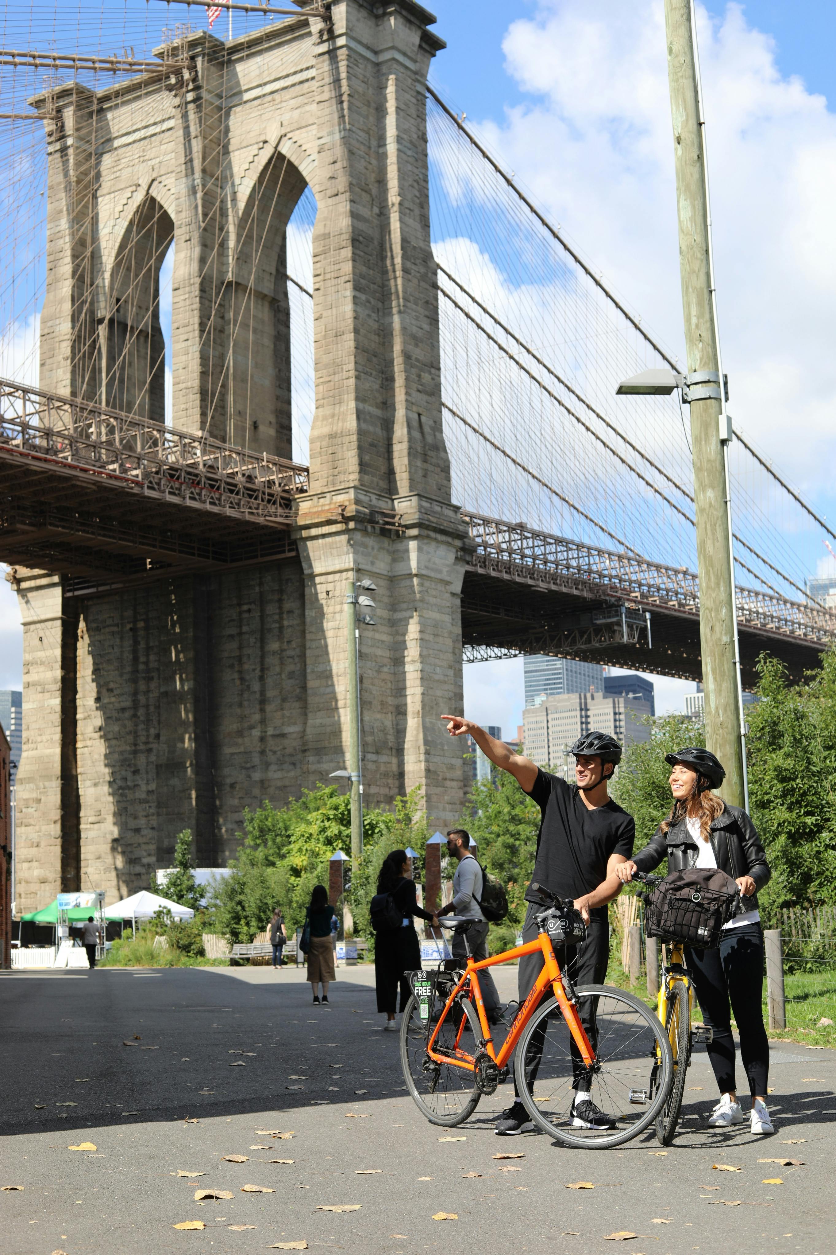 Un uomo e una donna in bicicletta si trovano di fronte al ponte di Brooklyn, con l'uomo che punta in avanti sotto un cielo sereno.