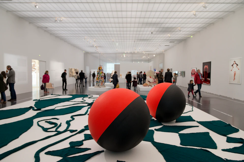 People exploring a spacious art gallery with large red and black spheres and various artworks displayed against white walls.