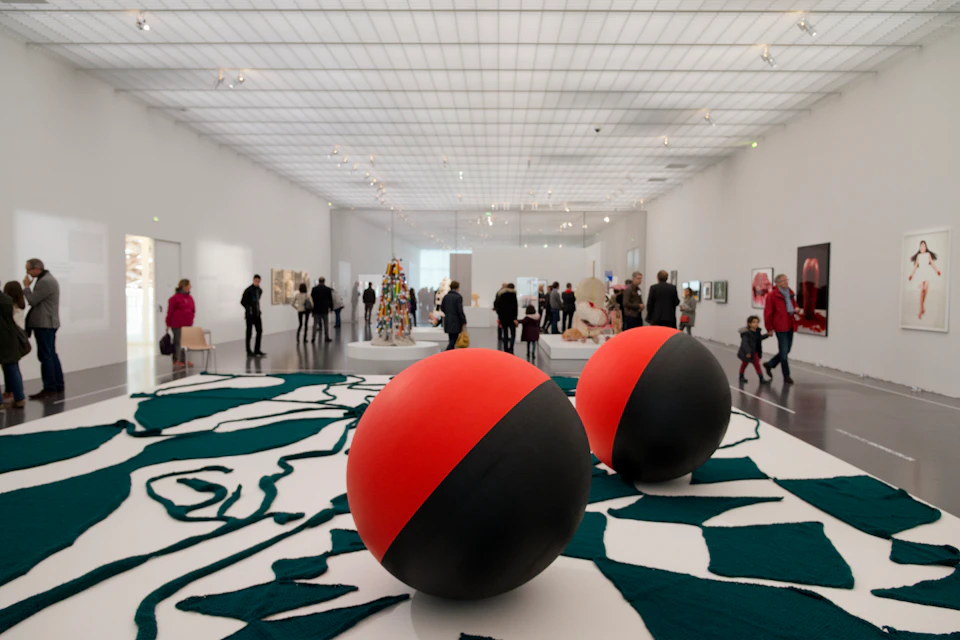 People exploring a spacious art gallery with large red and black spheres and various artworks displayed against white walls.