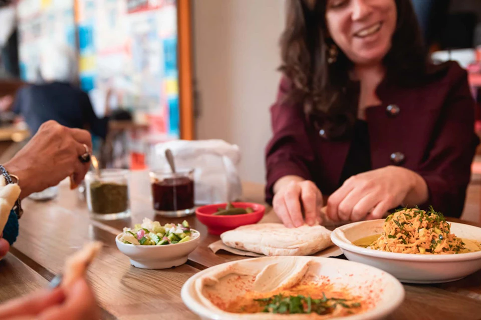 A smiling person preparing food at a table with dipping sauces, pita bread, and a plate of hummus in a relaxed setting.