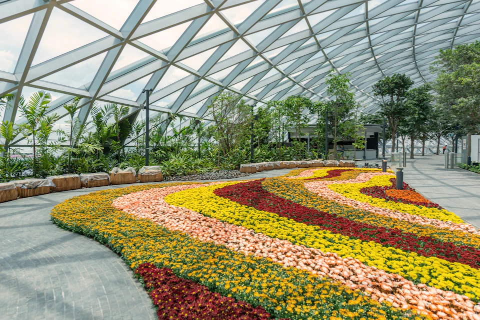 A spacious indoor garden with colorful flower beds arranged in wavy patterns, under a large glass dome roof.