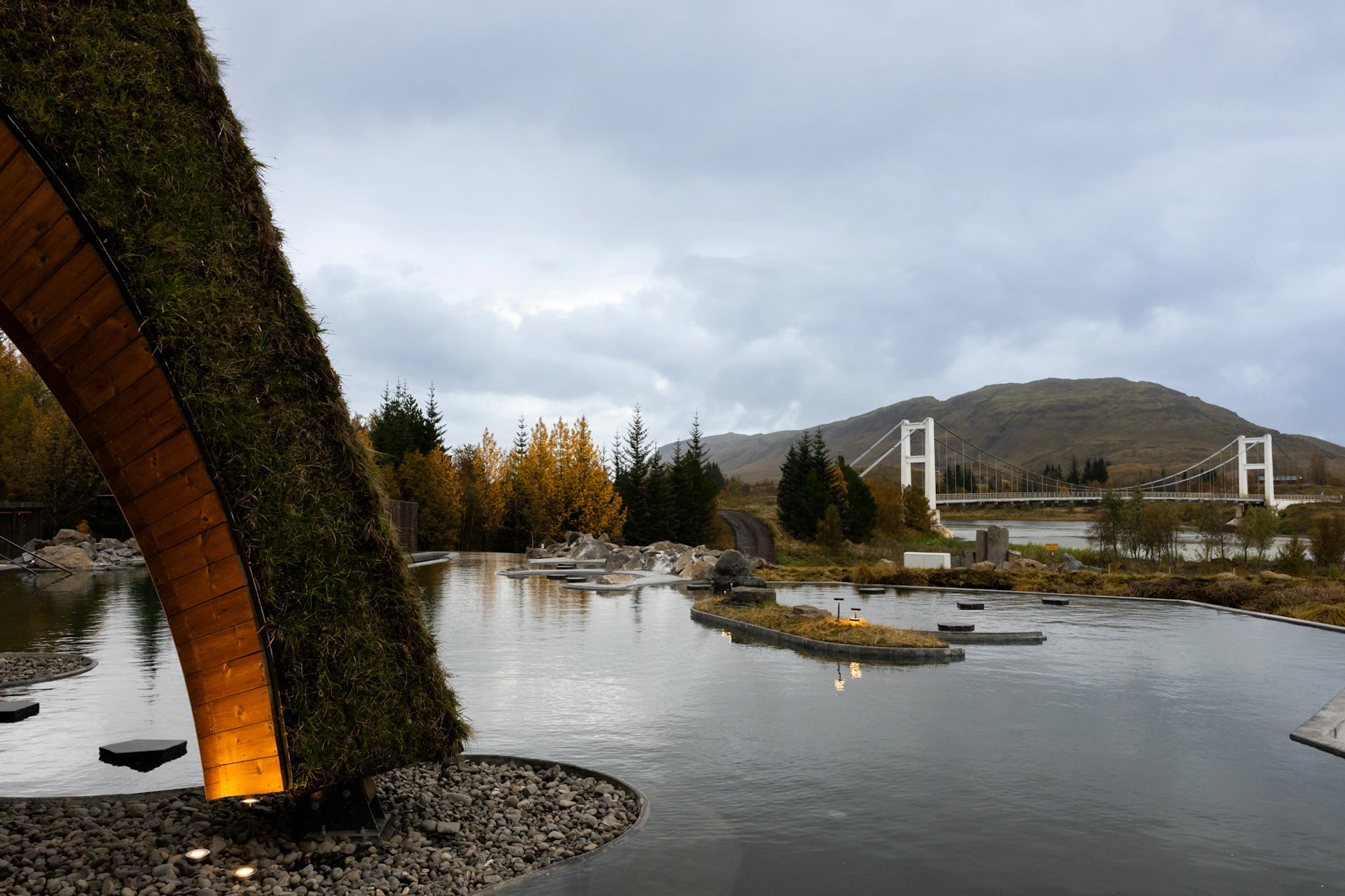 A serene outdoor scene featuring a triangular grass-roofed structure, a pond with small islands, trees, and a white bridge in the background.