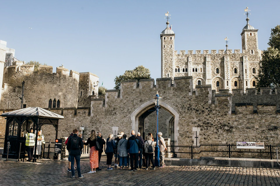 A group of tourists stands in front of a historical stone castle with a clock tower under a clear blue sky.
