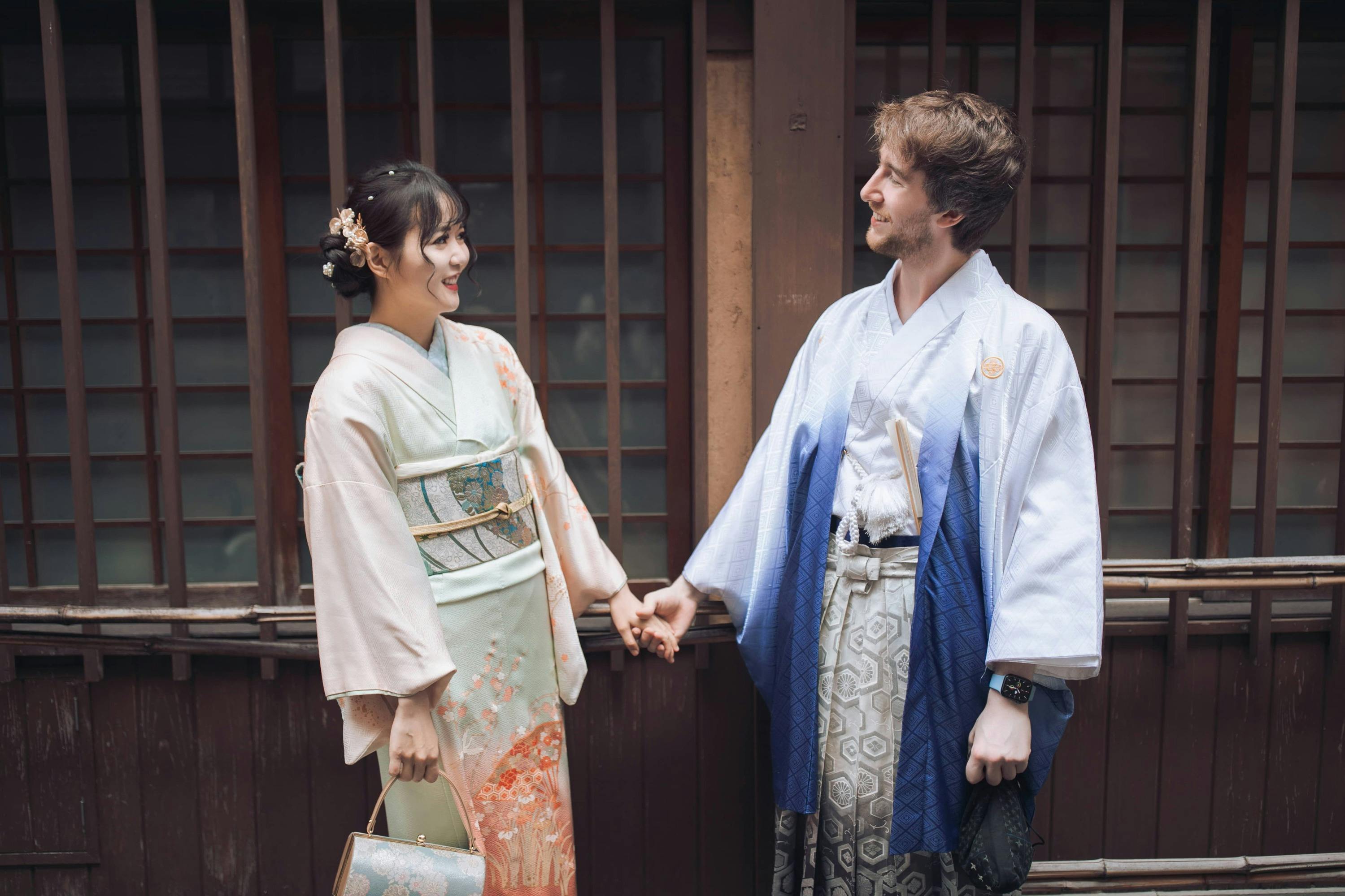A man and a woman wearing traditional Japanese attire hold hands and smile at each other outside a wooden building.