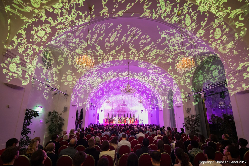 An audience watches a stage performance in a lavish hall with ornate floral light projections and chandeliers above.