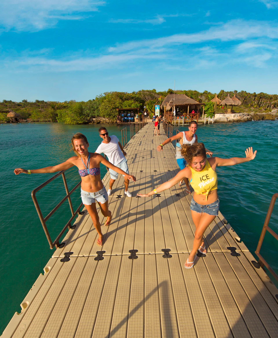 Four people joyfully running on a pier over blue water, with lush green landscape and thatched-roof huts in the background.