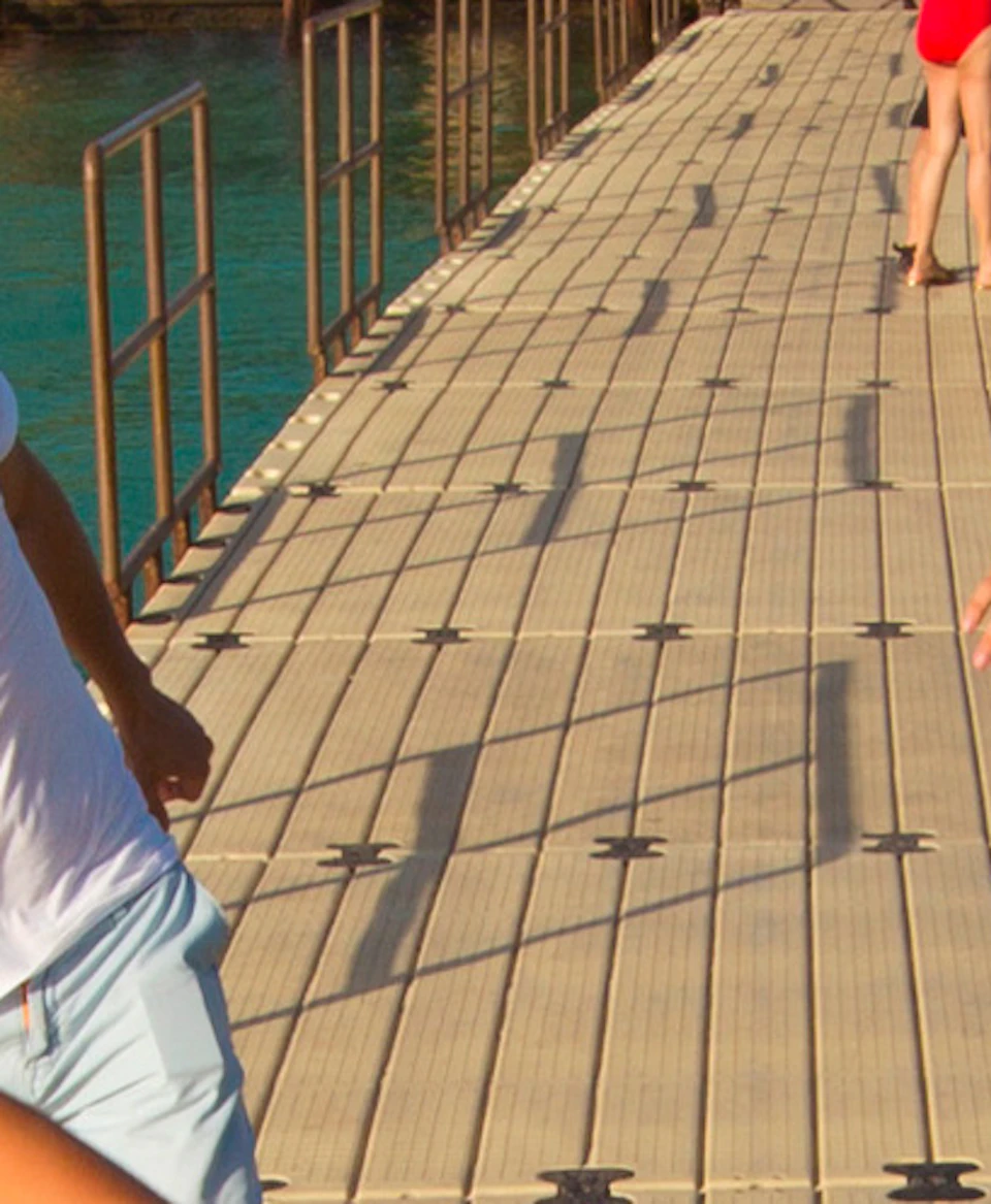 Four people joyfully running on a pier over blue water, with lush green landscape and thatched-roof huts in the background.
