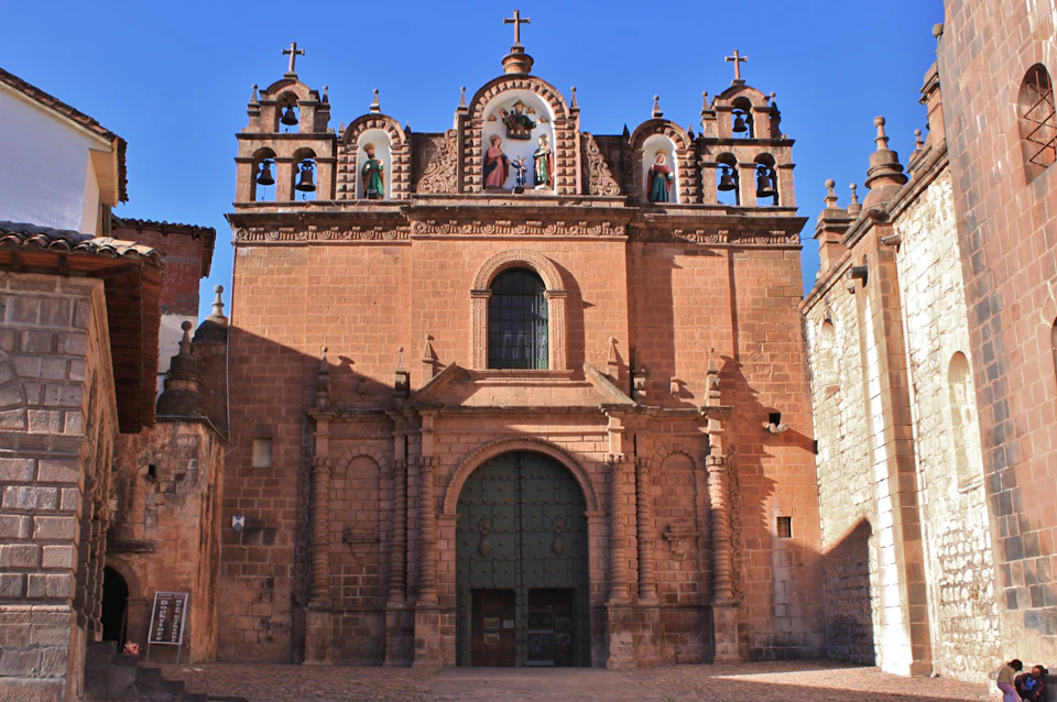 A stone church facade with religious statues, arched doorway, decorative windows, and crosses under a clear blue sky.