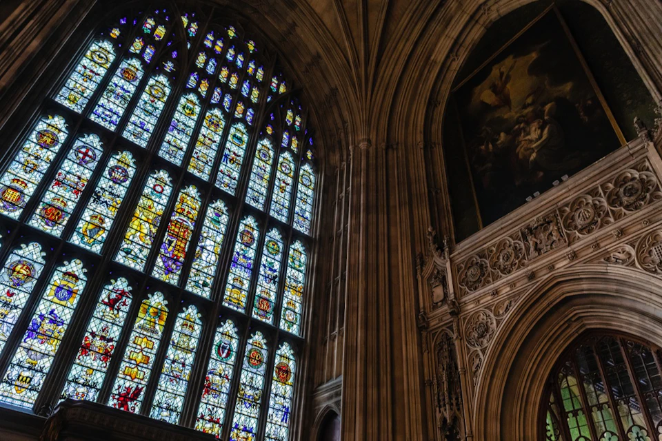 Large, arched stained glass window with intricate designs in a gothic-style building next to ornate stonework on the wall.
