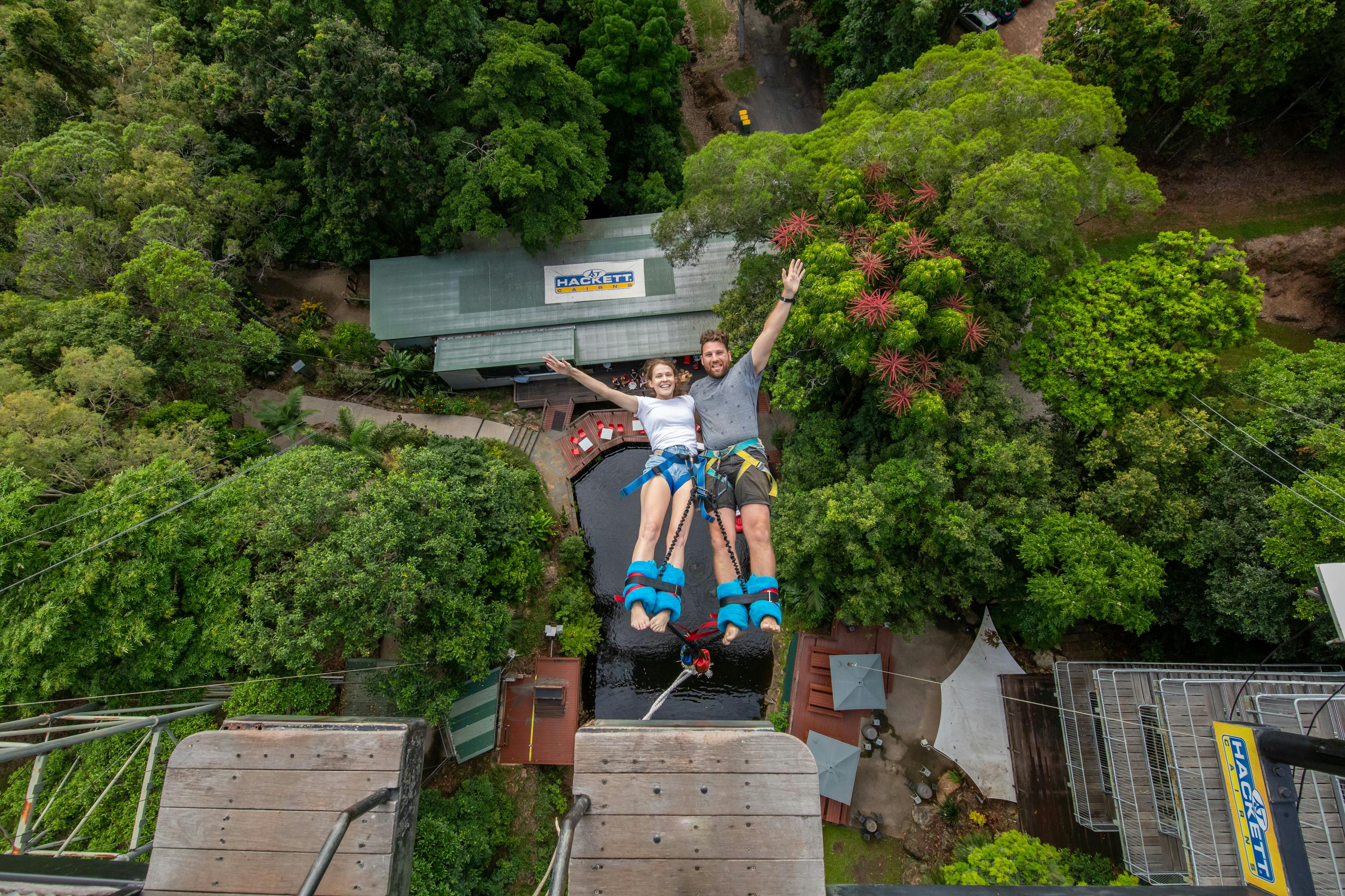 Two people are bungee jumping, descending towards a lush, green landscape with buildings and trees visible below.