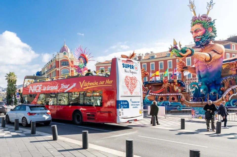 A red double-decker tour bus parked on a city street near colorful buildings and a festive carnival display.