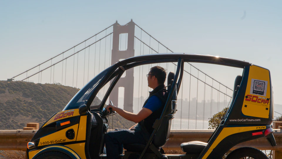 A person in a small enclosed vehicle with the Golden Gate Bridge visible in the background on a sunny day.