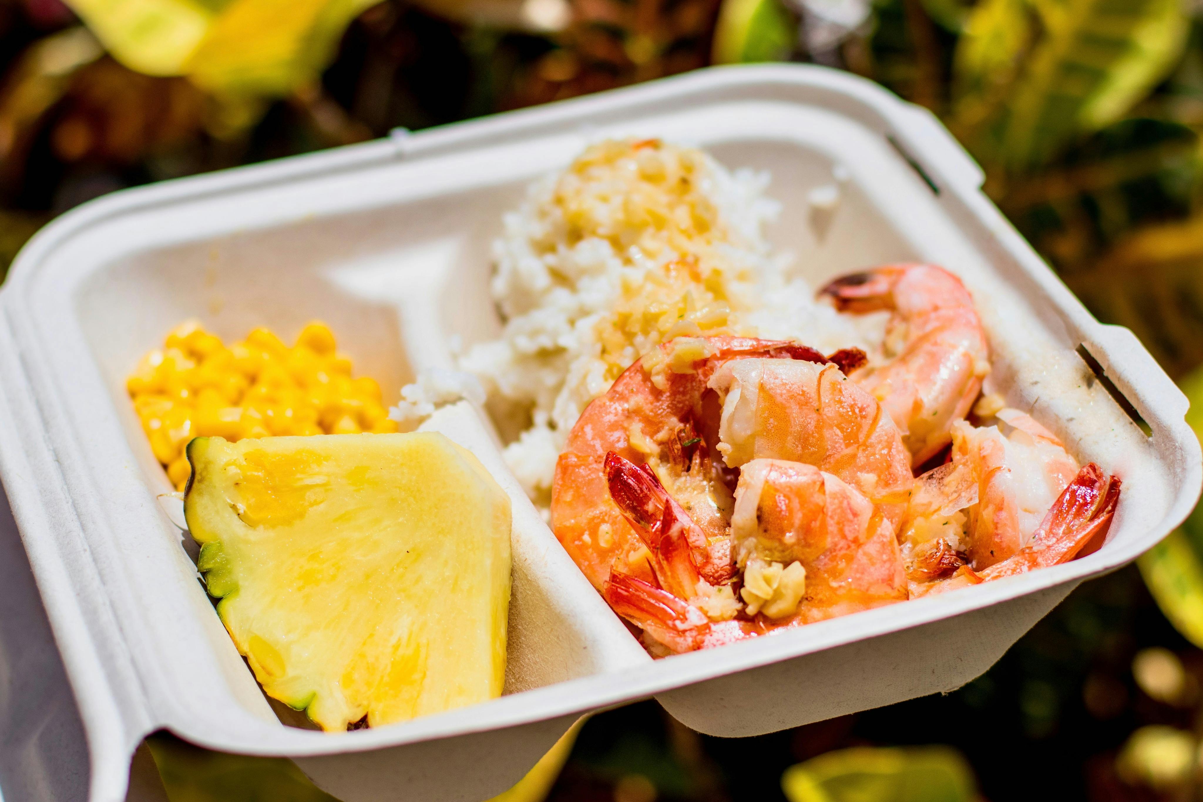 A takeout container with shrimp, rice, corn, and a slice of pineapple, placed against a blurred natural background.