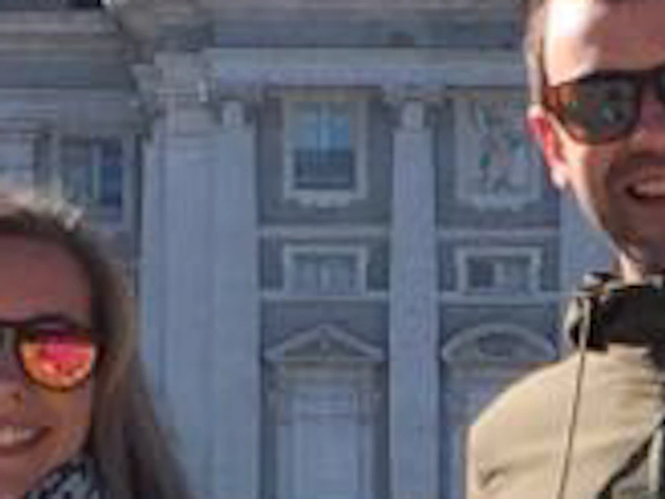 Two people with bicycles stand in front of a large building with a dome, surrounded by trees, on a clear sunny day.