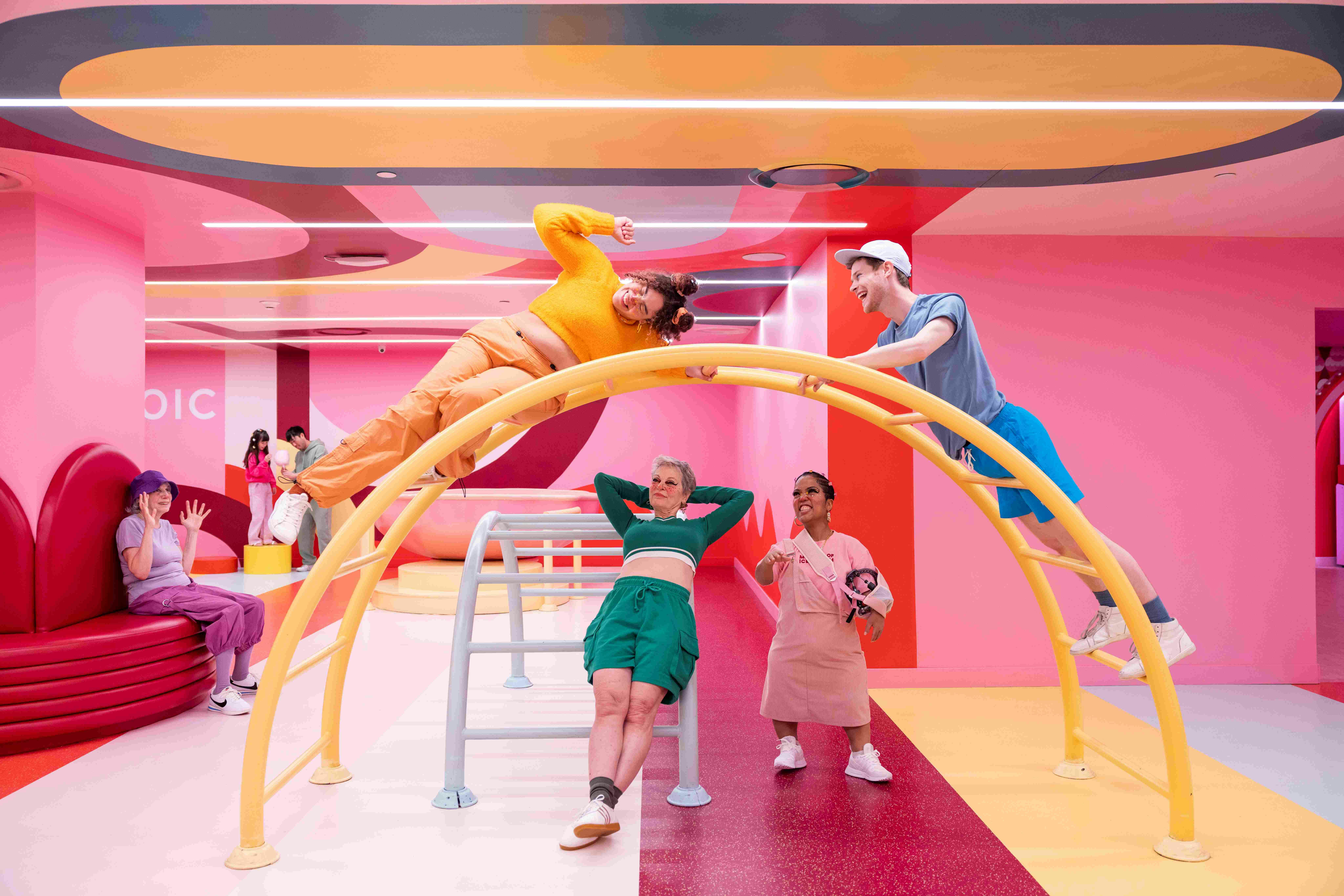 화려한 색상의 의상을 입은 네 명의 사람이 생생하게 칠해진 장난기 넘치는 방에서 노란색 아치형 구조물과 상호 작용합니다.