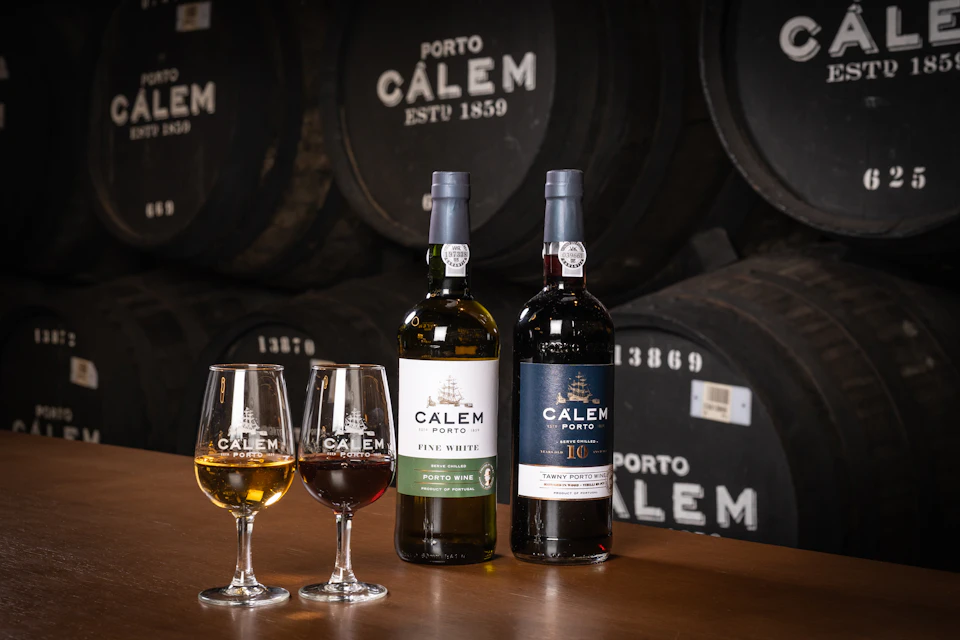 Two bottles of Calem Porto wine, glasses of white and red wine, and barrels in the background.