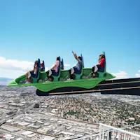 Vier mensen in een groene achtbaan die zich uitstrekt over de skyline van een stad met een helderblauwe lucht op de achtergrond.