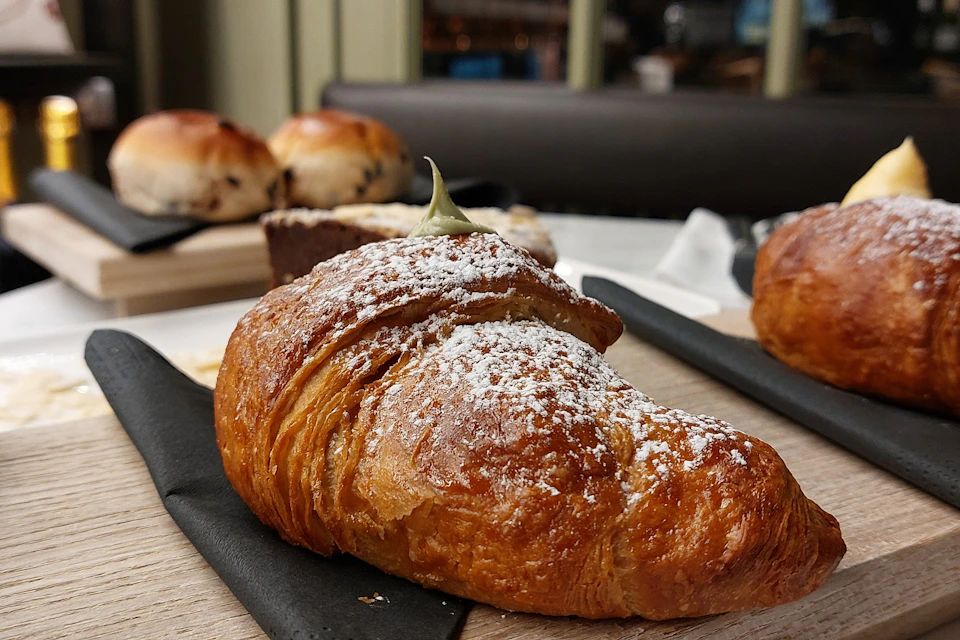 A close-up of a croissant dusted with powdered sugar, with other pastries blurred in the background.