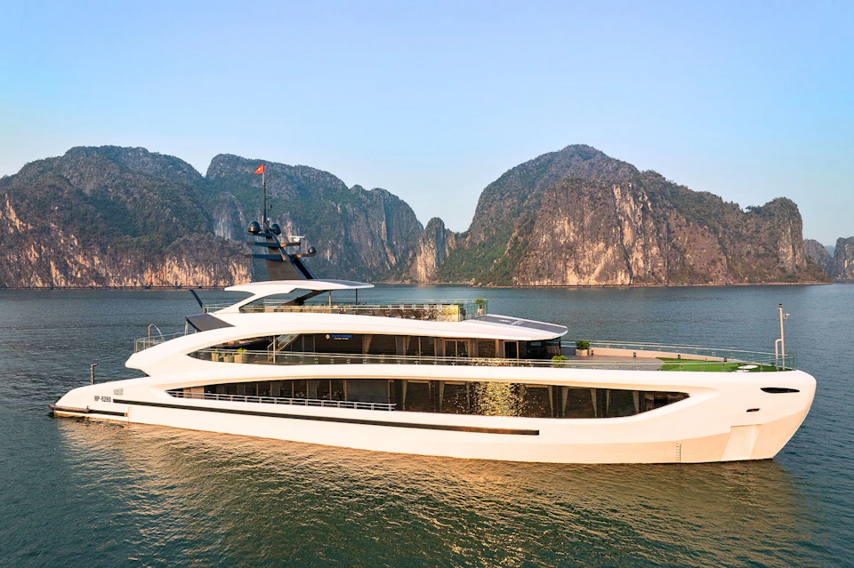Luxury yacht on tranquil water with green mountainous cliffs in the background under a clear, blue sky.