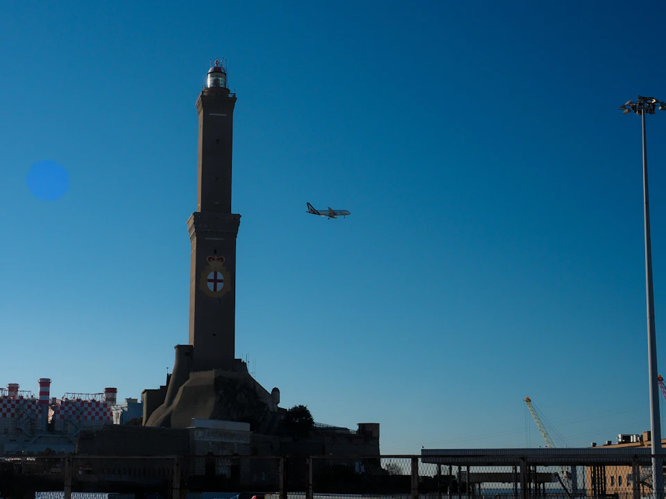 A tall lighthouse with a cross emblem, an airplane flying in the blue sky, and industrial structures below.