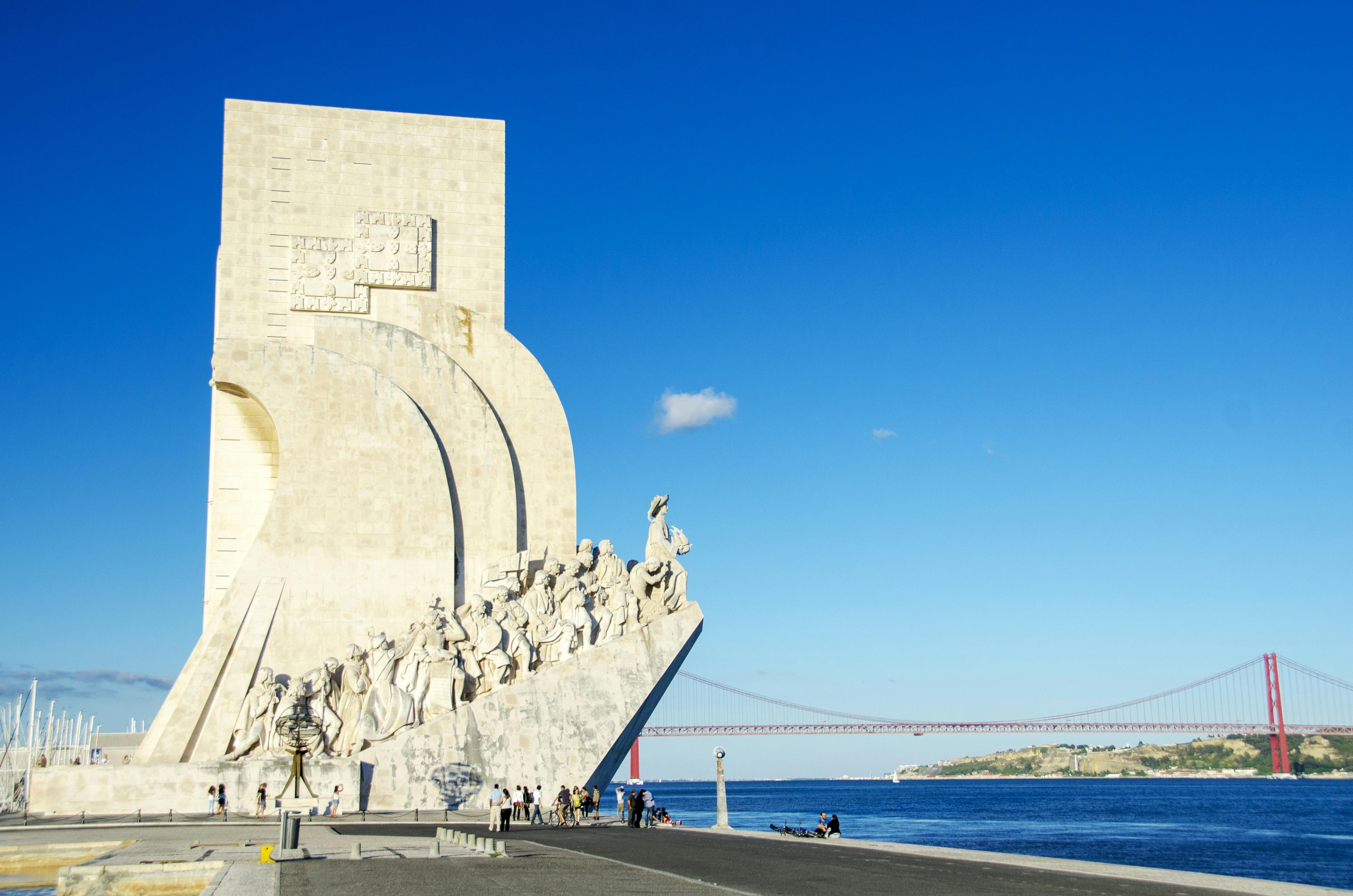 Monument with sculptures of figures overlooking a river, with a distant bridge and clear blue sky. A few people sit by the water.