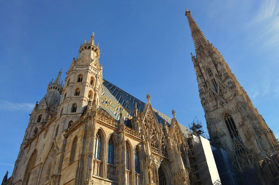 Gothic cathedral with detailed spires and ornate windows against a clear blue sky. One spire is under construction.