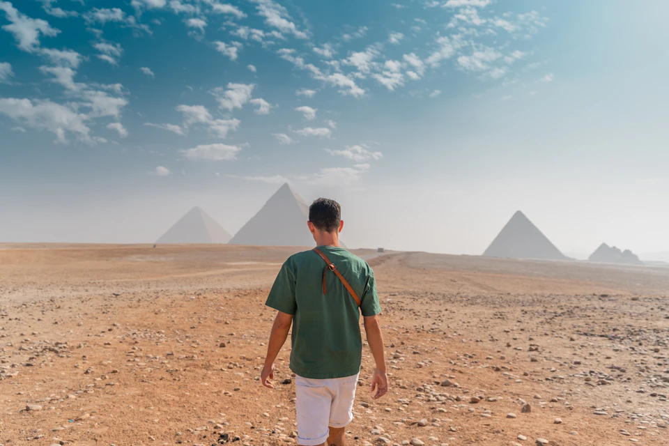 A person in a green shirt and white shorts walking toward the pyramids in a desert under a partly cloudy sky.