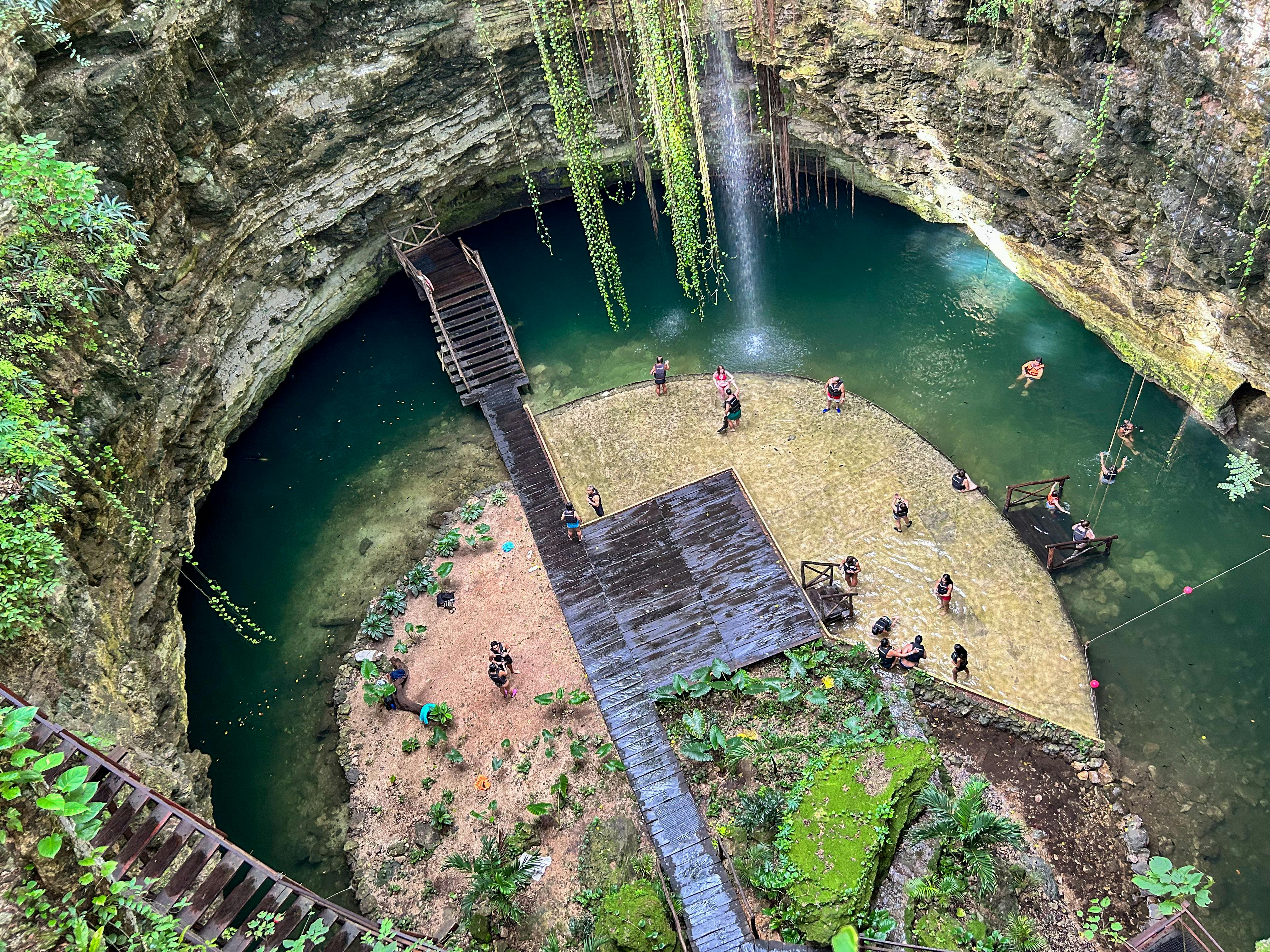 Persones nedant i de peu sobre plataformes en un cenote circular i rocós amb cascada, vegetació exuberant i passarel·les de fusta.