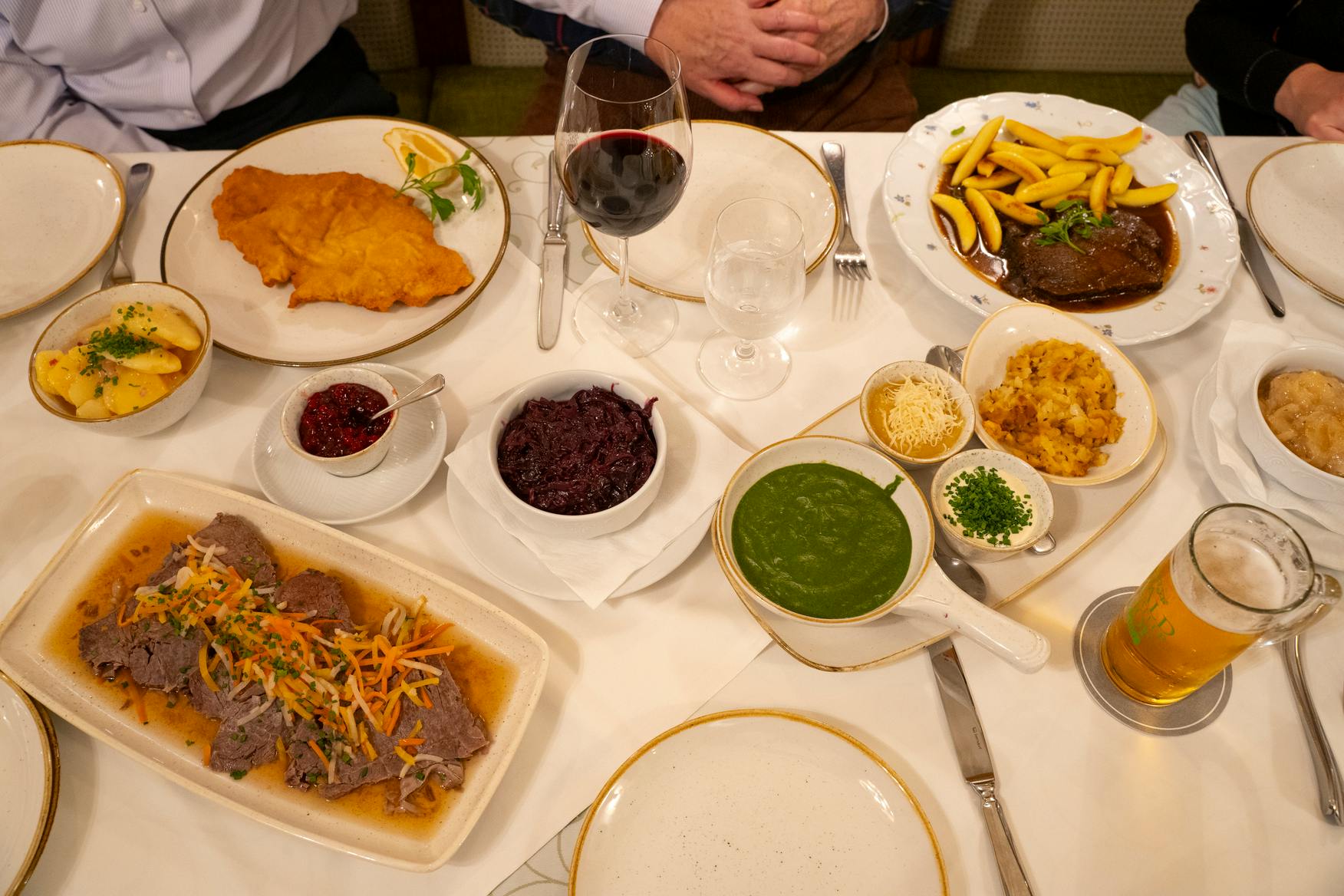 Uma mesa posta com vários pratos, incluindo schnitzel, legumes, molhos e copos de vinho e água, com duas pessoas sentadas.