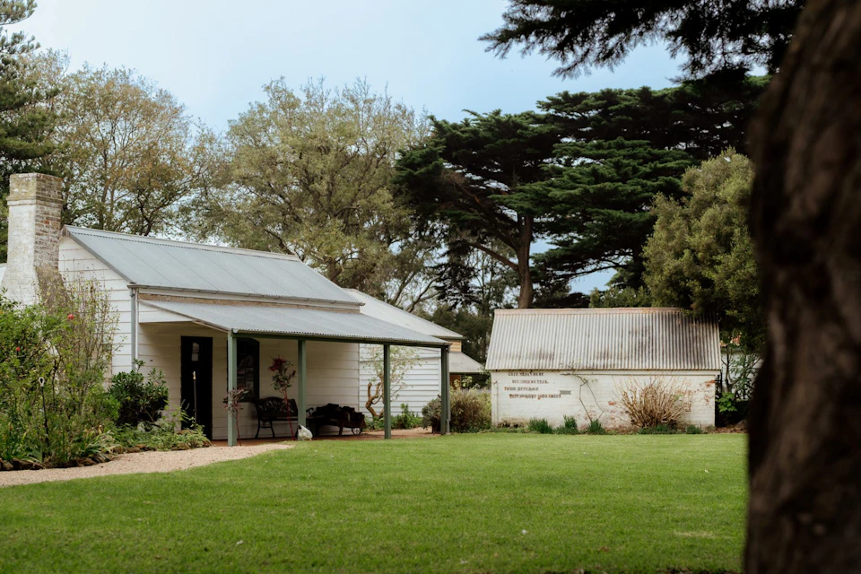 Single-story house with a covered porch next to a small shed. Green lawn and large trees in the background.