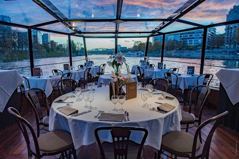 A riverside restaurant with a glass ceiling and large windows. Tables are set with white tablecloths, glassware, and floral centrepieces.