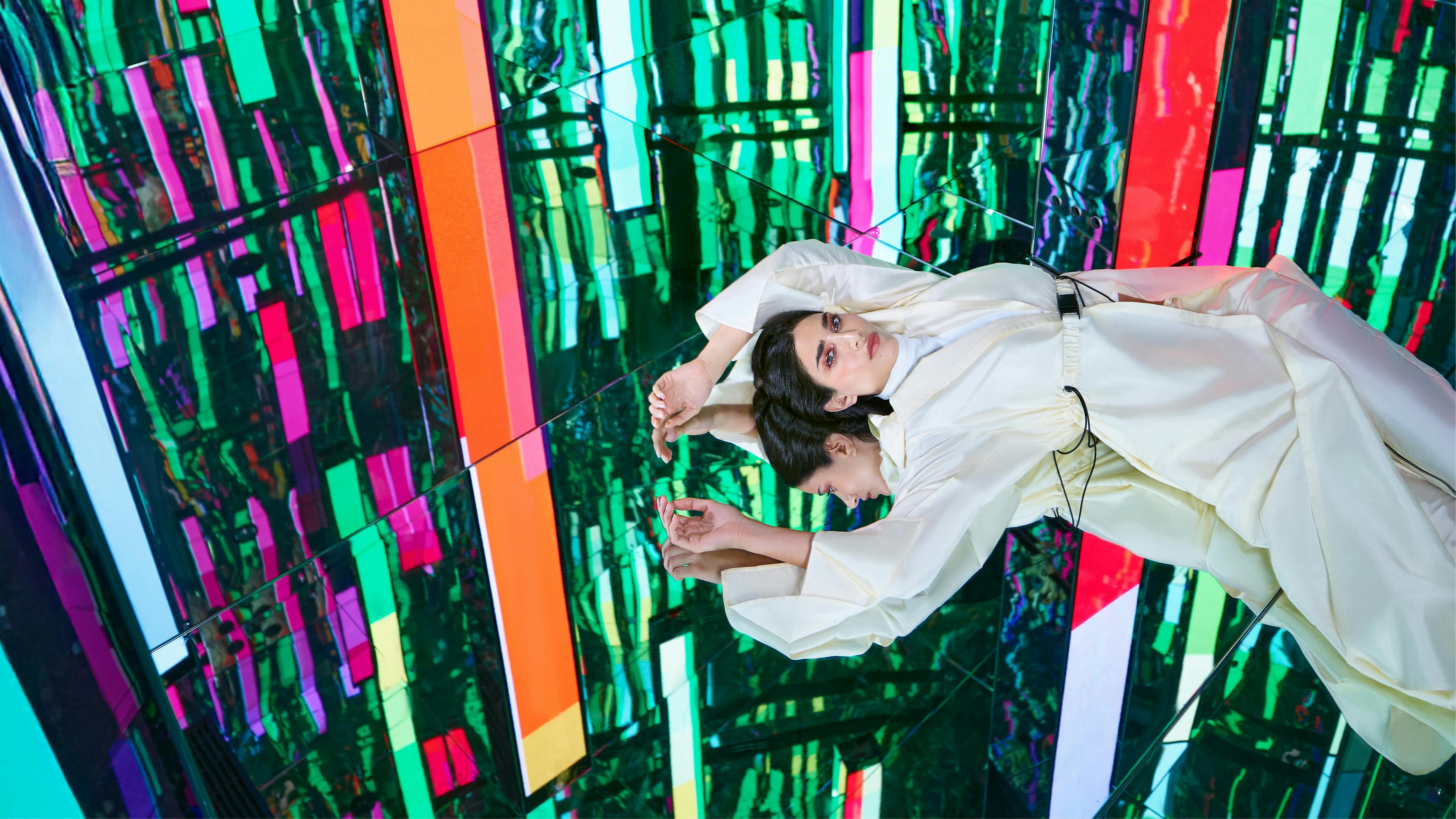 Μια γυναίκα με λευκό ντύσιμο βρίσκεται σε μια ανακλαστική επιφάνεια με πολύχρωμα, αφηρημένα μοτίβα φωτός στο παρασκήνιο.