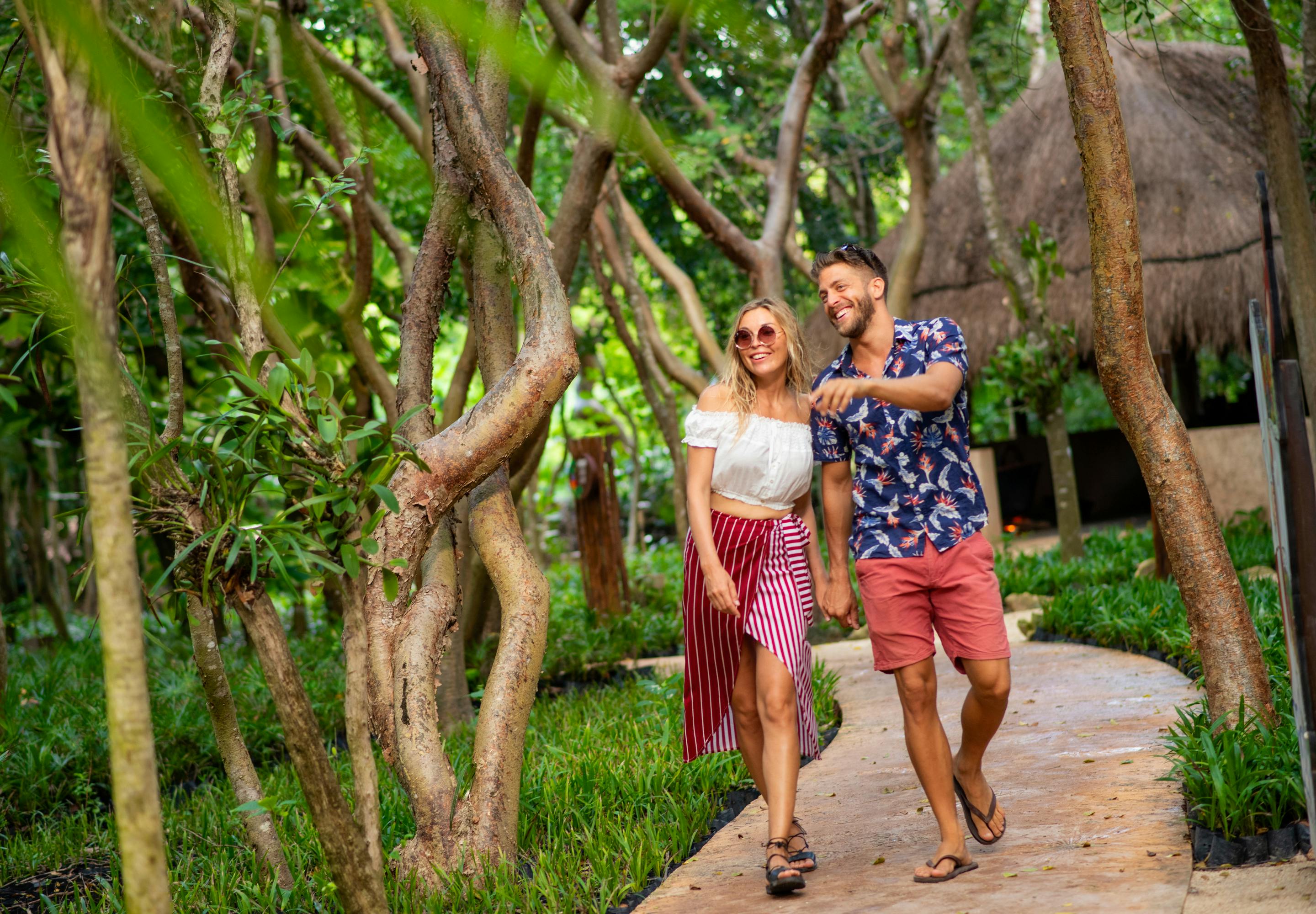 Un couple souriant marche main dans la main sur un chemin bordé d'arbres, près d'une cabane au toit de chaume, entourée d'une végétation luxuriante.