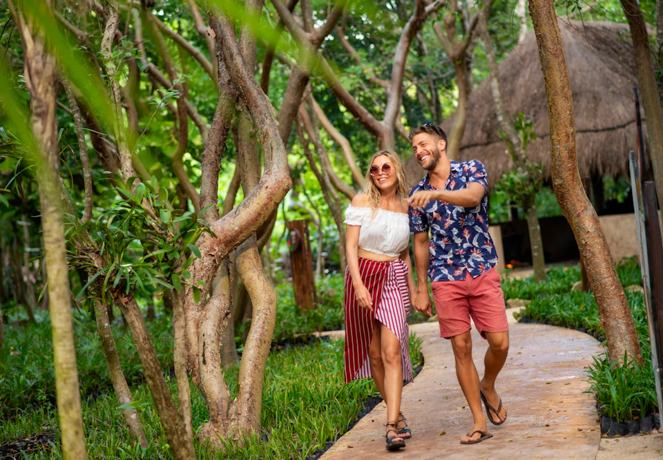 A smiling couple walks hand-in-hand down a tree-lined path near a thatched-roof hut, surrounded by lush greenery.
