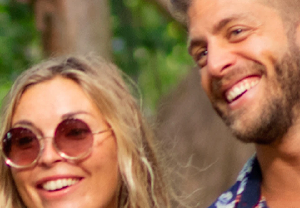 A smiling couple walks hand-in-hand down a tree-lined path near a thatched-roof hut, surrounded by lush greenery.