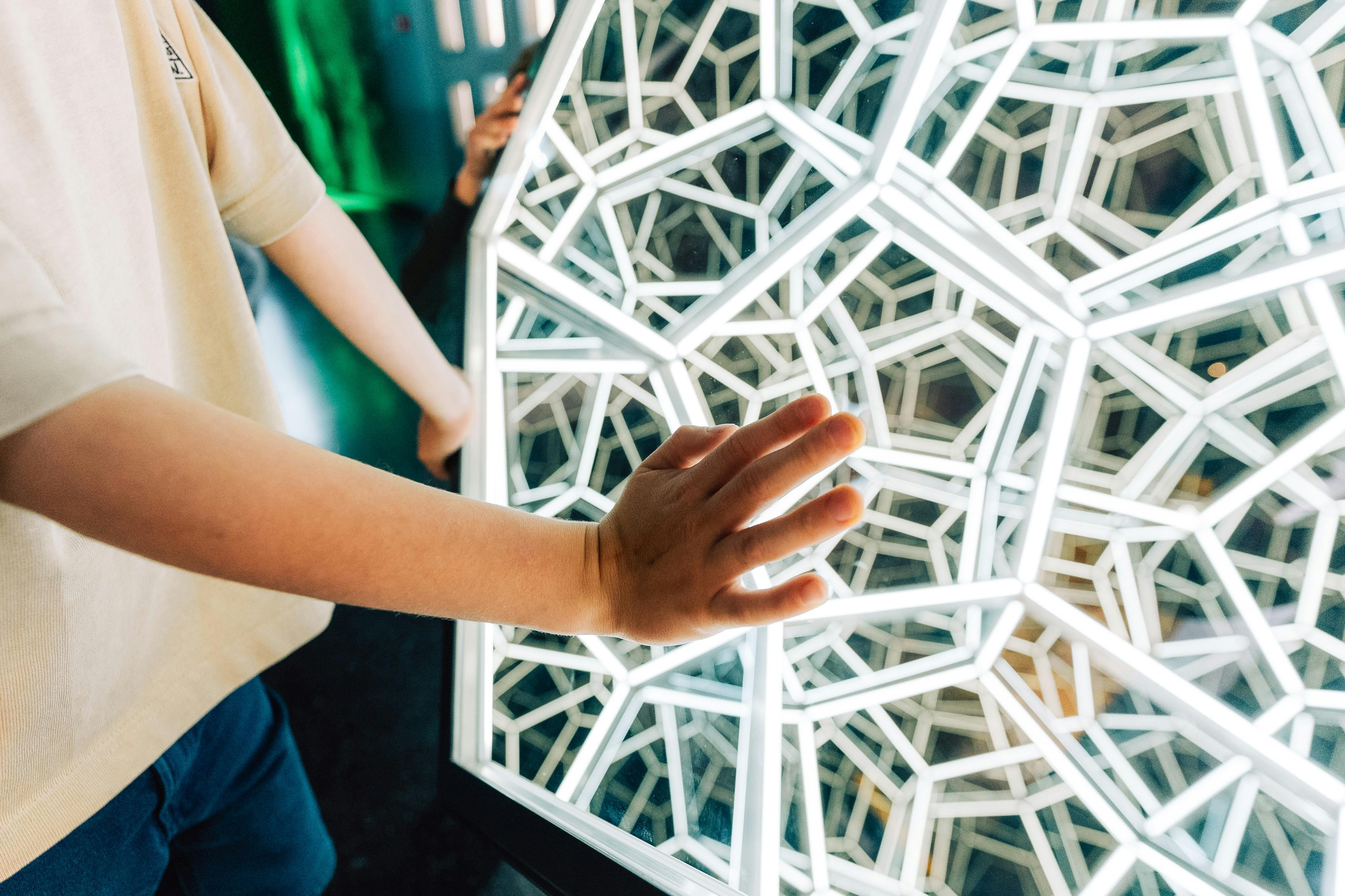 A child’s hand touches a geometric, illuminated structure composed of interconnected, glowing lines. Another person's arm is partially visible.