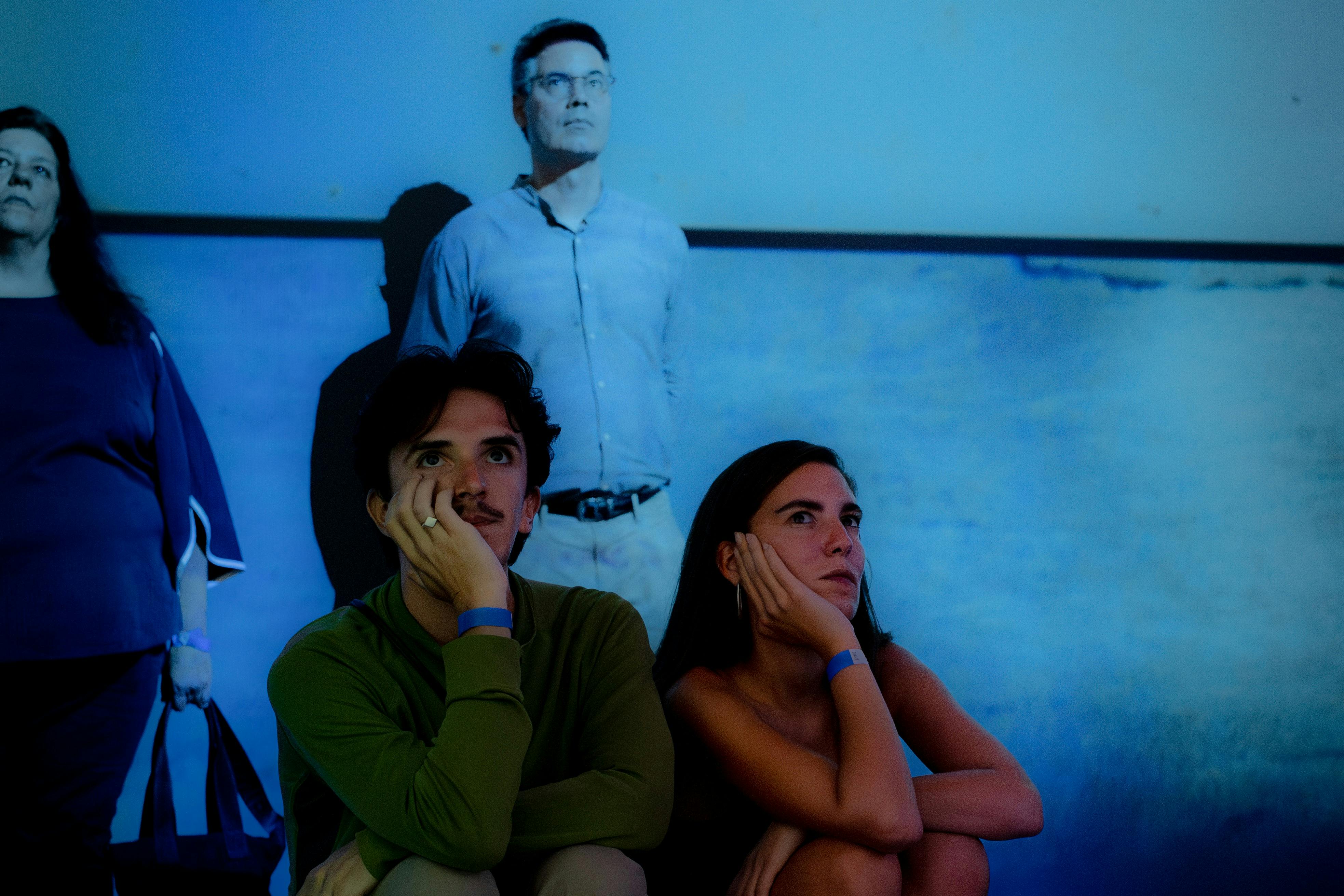 Čtyři lidé v tlumeně osvětlené místnosti s modrým osvětlením; dva sedí v popředí a zamyšleně vzhlížejí, dva stojí v pozadí.