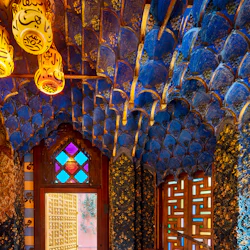 Utsmyckat rum med tak i blått och guld, glasmålning, mönstrade kakelväggar och en geometrisk trädörr med färgat glas.